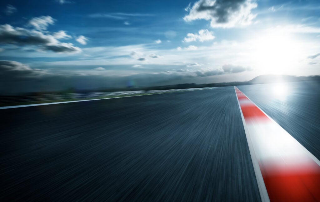 Blurred view of a race track at high speed under a dramatic sky, with a red and white curb line leading into the distance and the sun shining brightly on the horizon.