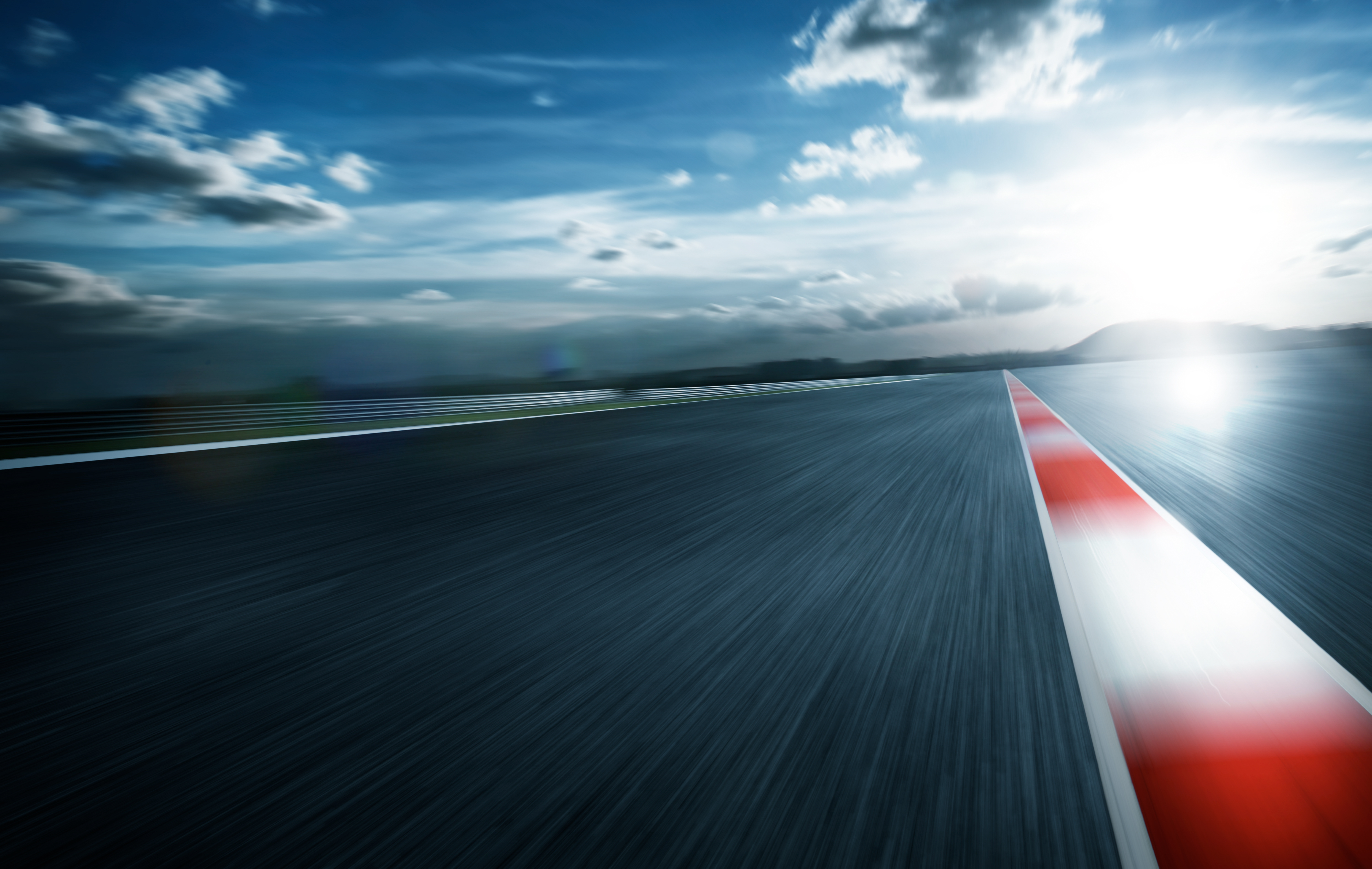 Blurred view of a race track at high speed under a dramatic sky, with a red and white curb line leading into the distance and the sun shining brightly on the horizon.