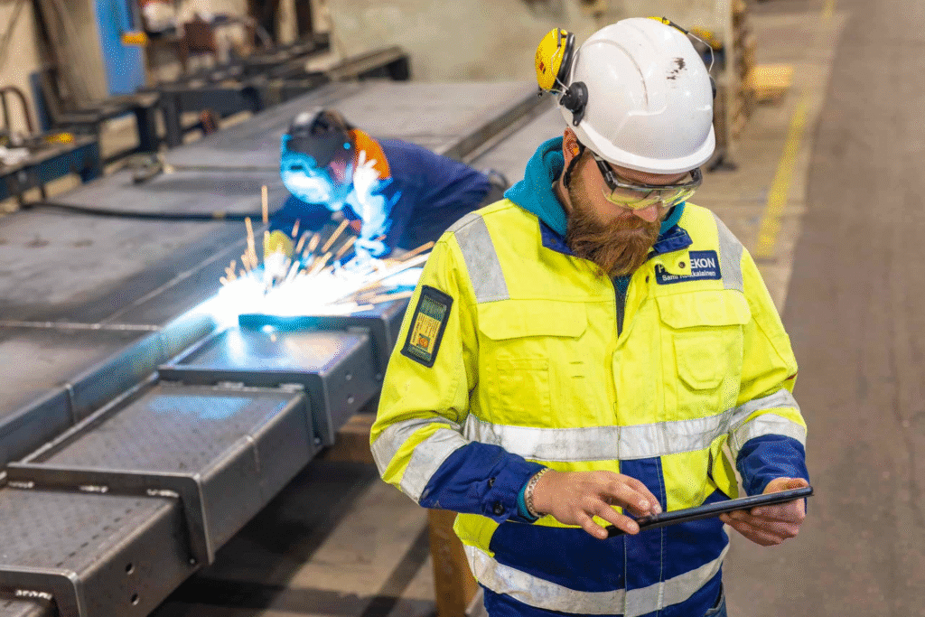 Engineer in high-visibility safety gear using a tablet on a factory floor while a colleague welds metal components in the background, illustrating digital tools in industrial manufacturing.