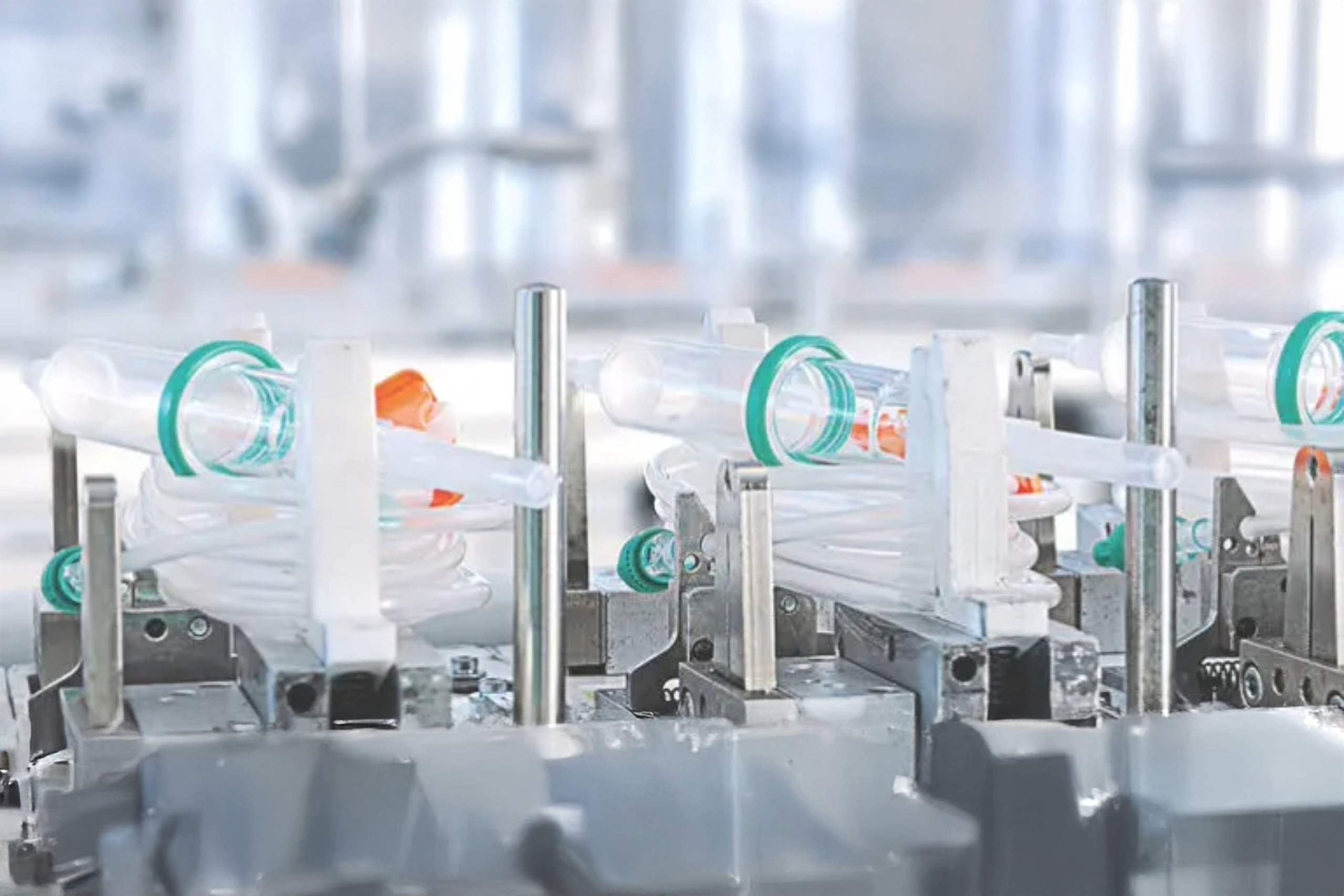 Automated medical device manufacturing line assembling clear plastic tubing and connectors, including green and orange components, in a sterile, high-tech environment.